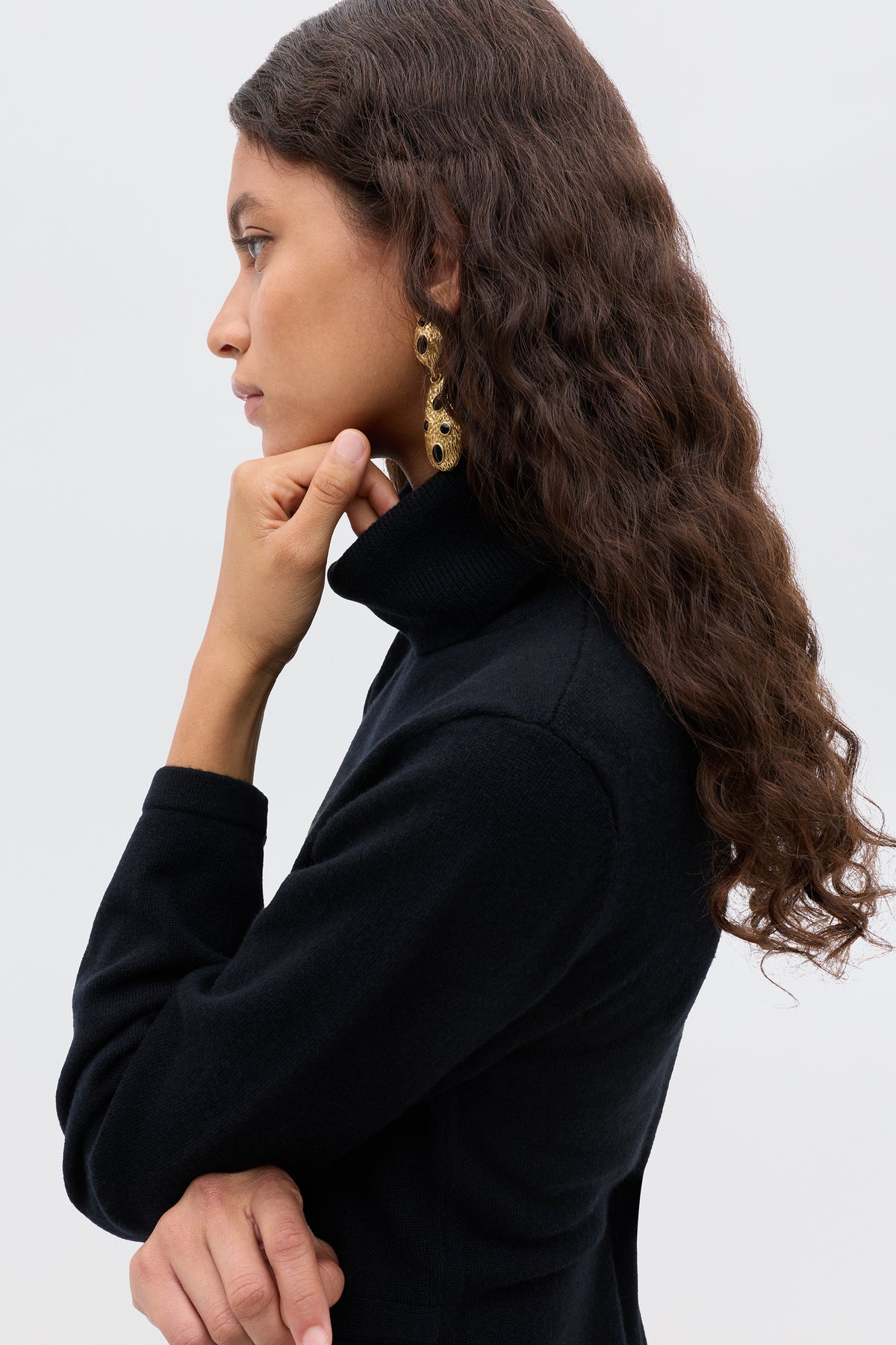 A woman with long, wavy brown hair wears the Aly - Black turtleneck and gold statement earrings, standing in profile with her hand on her chin against a plain light background.