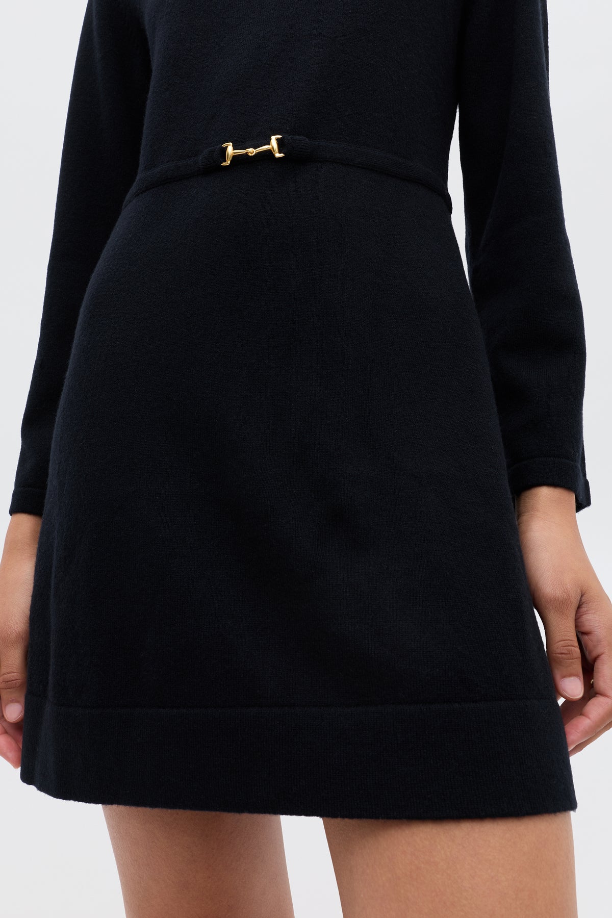 A person wearing the Aly - Black long-sleeve winter LBD with a fitted waist and a small gold buckle detail, shown from shoulders to mid-thigh.