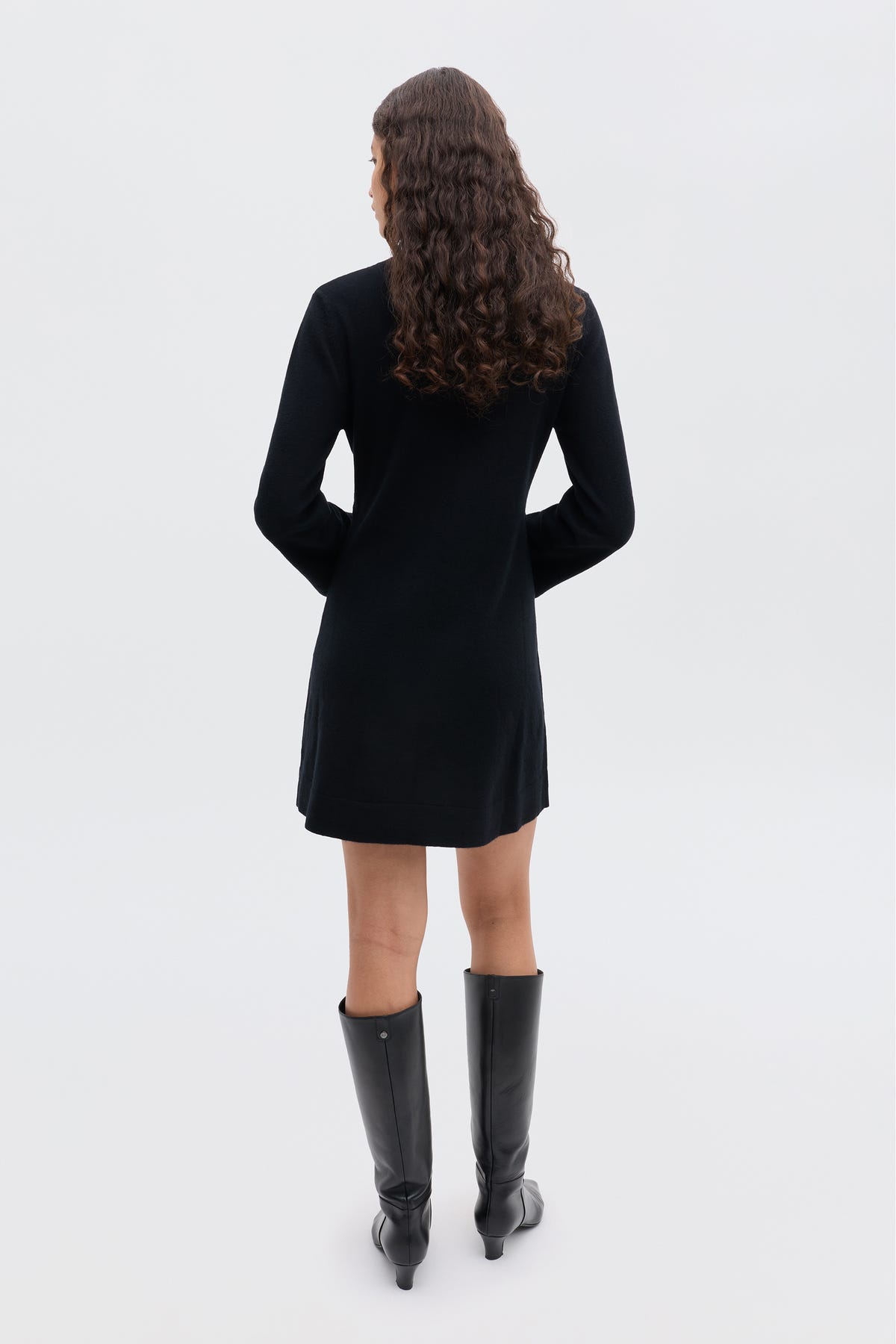 A person with long, curly brown hair stands with their back to the camera, wearing the Aly - Black winter LBD and knee-high black boots against a plain light background.