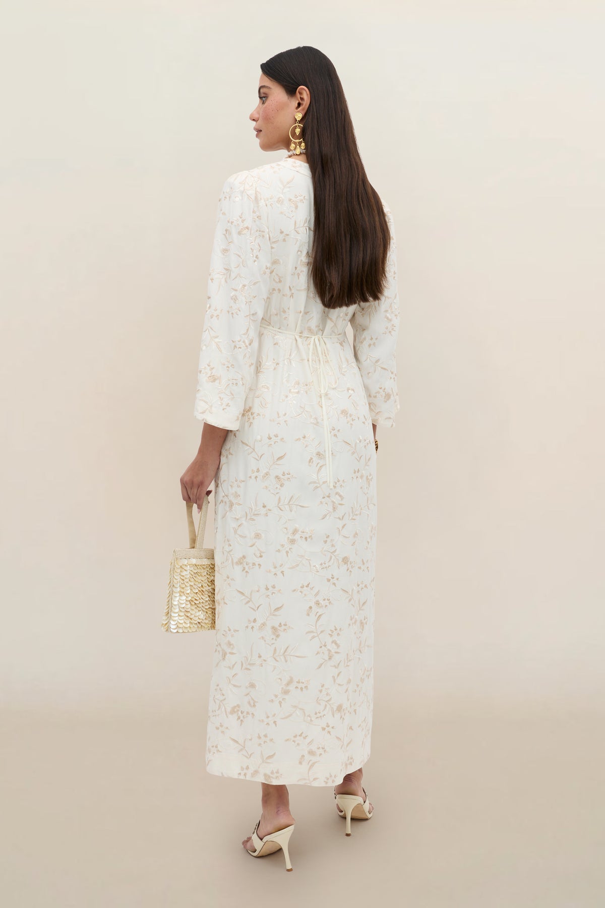 Woman wearing the Dahlia - Botanical Embroidery Pearl dress, a long white vintage-inspired gown featuring embroidered floral designs and long sleeves. She holds a woven handbag, pairs the look with gold earrings and cream heels, and stands against a plain beige background, facing away from the camera.
