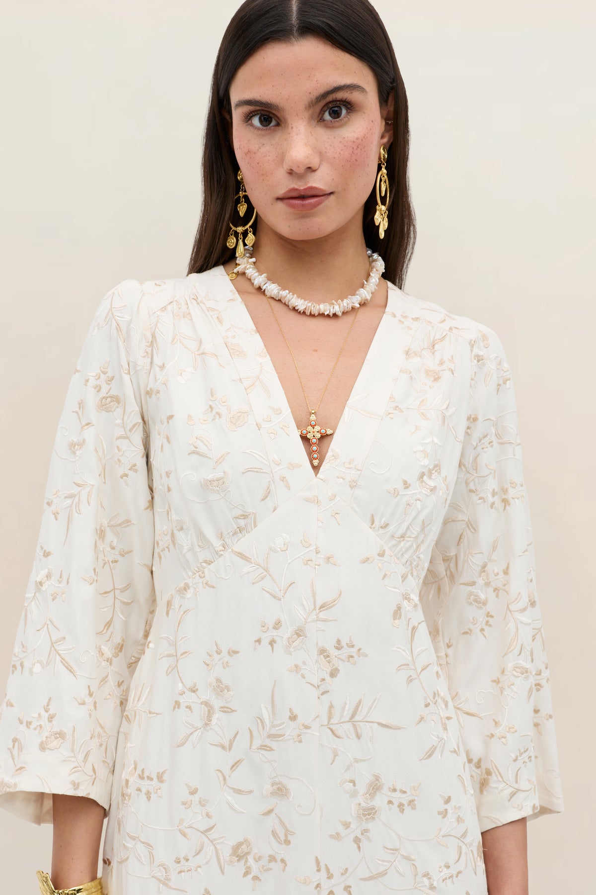 Woman with long dark hair wearing the “Dahlia - Botanical Embroidery Pearl” dress, a cream vintage-inspired piece with subtle floral embroidery, styled with gold earrings, layered necklaces, and a delicate beaded choker, standing against a plain beige background.