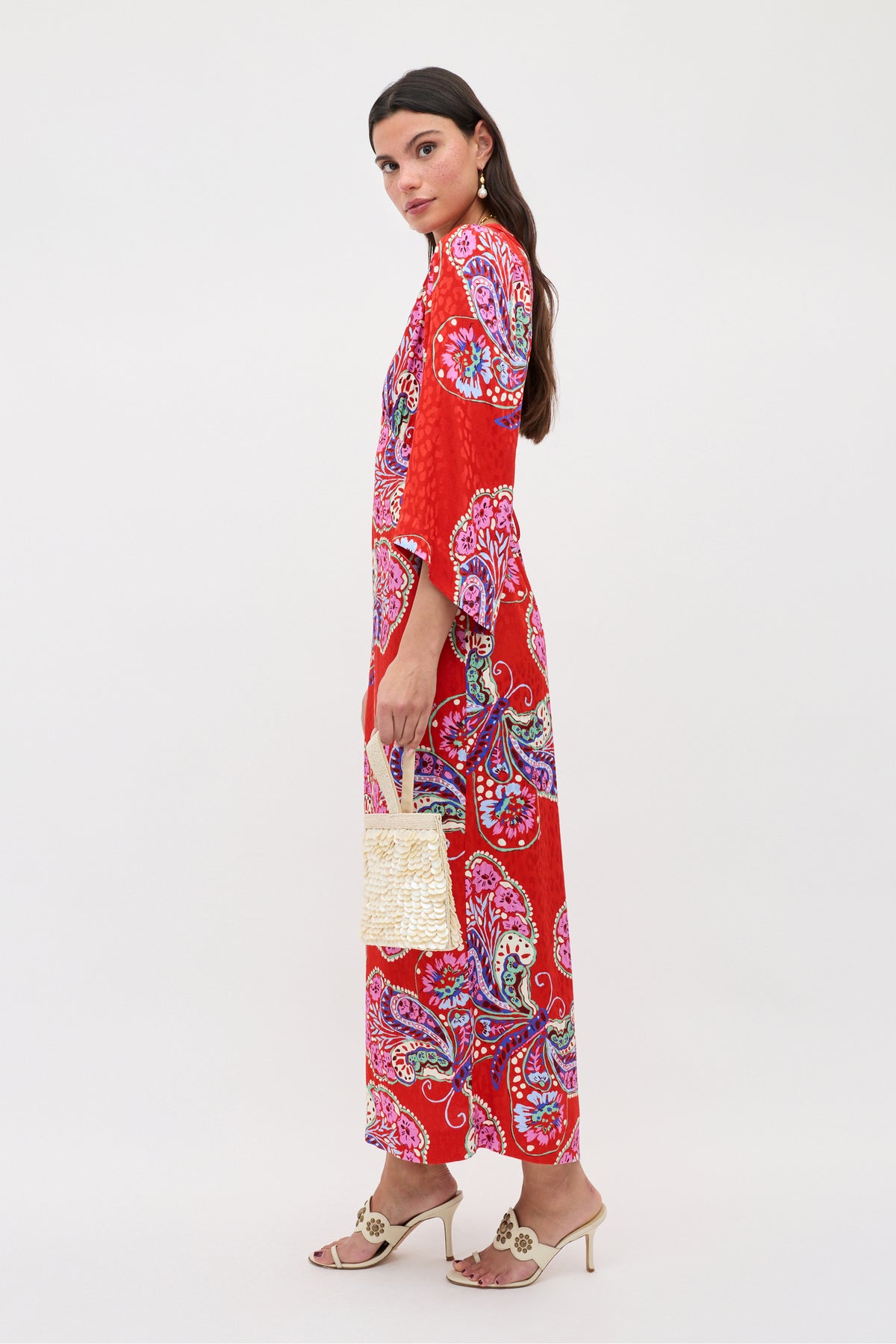 Wearing the Dahlia - Butterfly Bloom Jacquard Red dress, a woman with long dark hair stands sideways, holding a woven handbag and wearing open-toe heeled sandals and neutral earrings against a plain background.