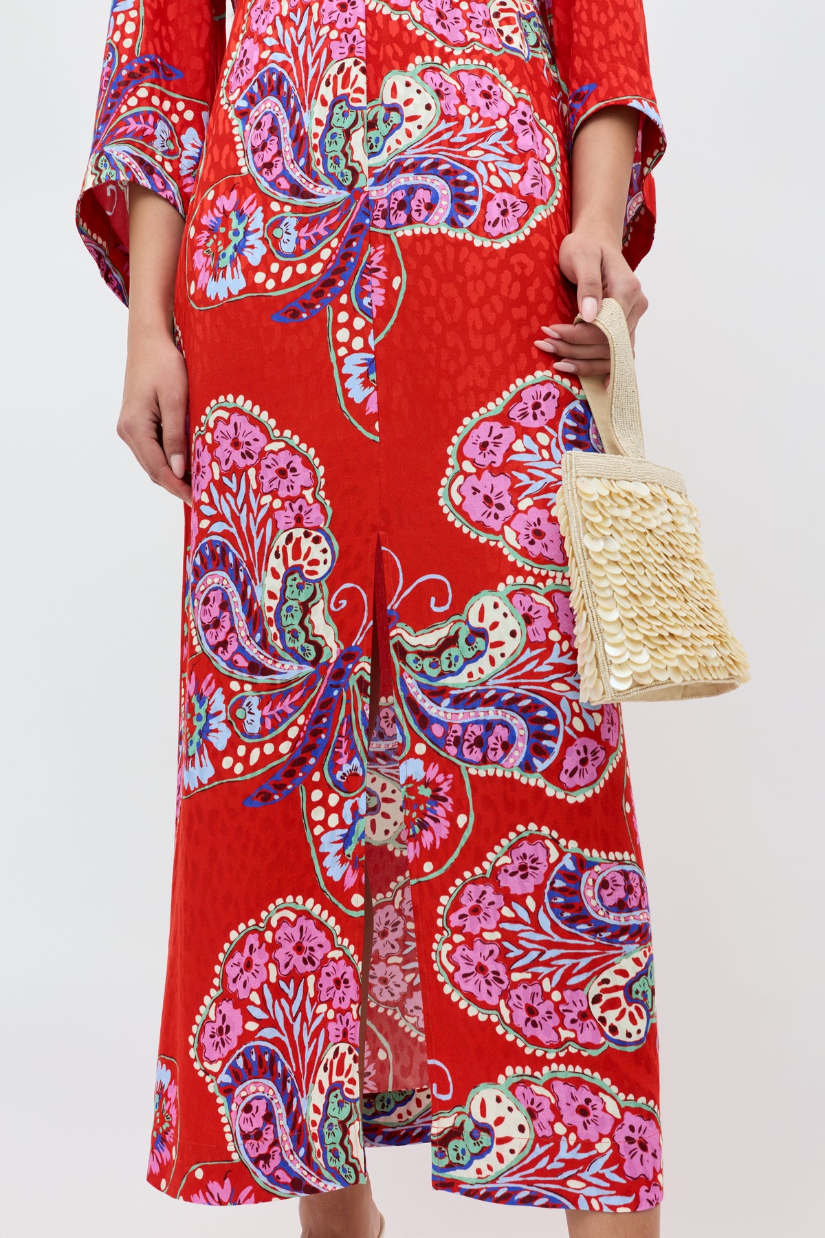 Wearing the Dahlia - Butterfly Bloom Jacquard Red dress with vibrant floral and paisley patterns, a person holds a textured beige handbag. Only their body from shoulders to feet is visible against a plain background.