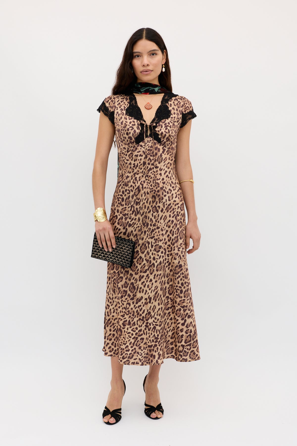 A woman poses against a white background wearing the Amari - Mila Leopard Brown short-sleeve midi dress with lace trim, paired with a black clutch, black heels, gold jewelry, and a black scarf around her neck.
