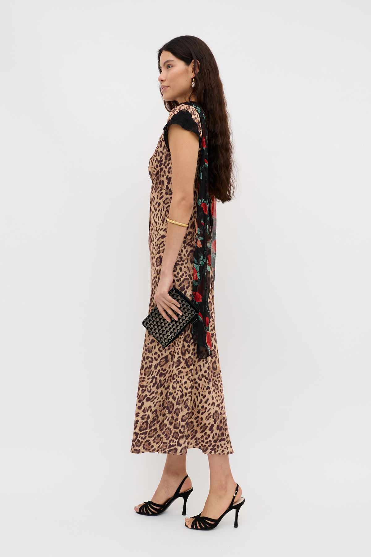 A woman stands in profile against a white background wearing the Amari - Mila Leopard Brown bias cut dress, black heeled sandals, a black floral scarf over one shoulder, and holding a black clutch.