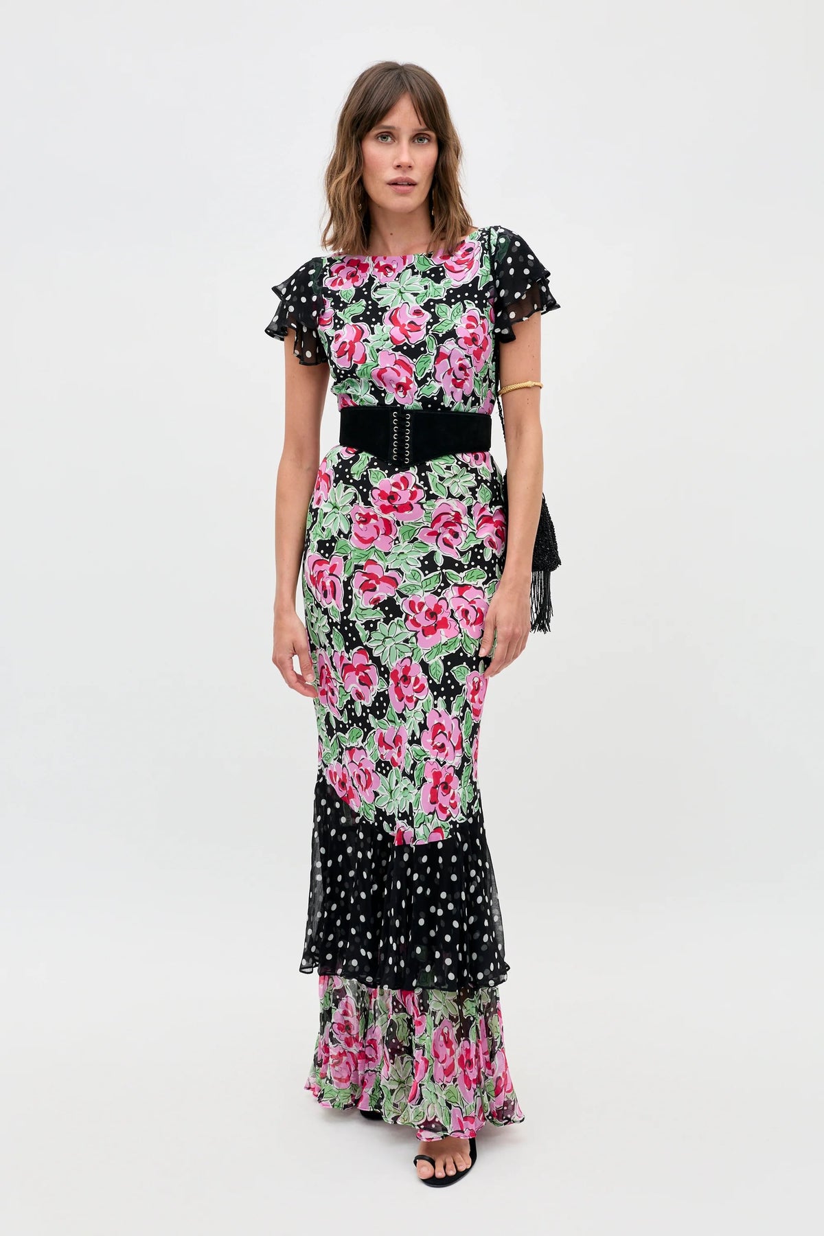 A woman models the Ordinia - Rose Dot Mix Black, a floor-length dress with a vibrant pink floral pattern and black-and-white polka dot ruffle sleeves, waistband, and hem. She has loose hair, black open-toe heels, and stands against a plain white background.