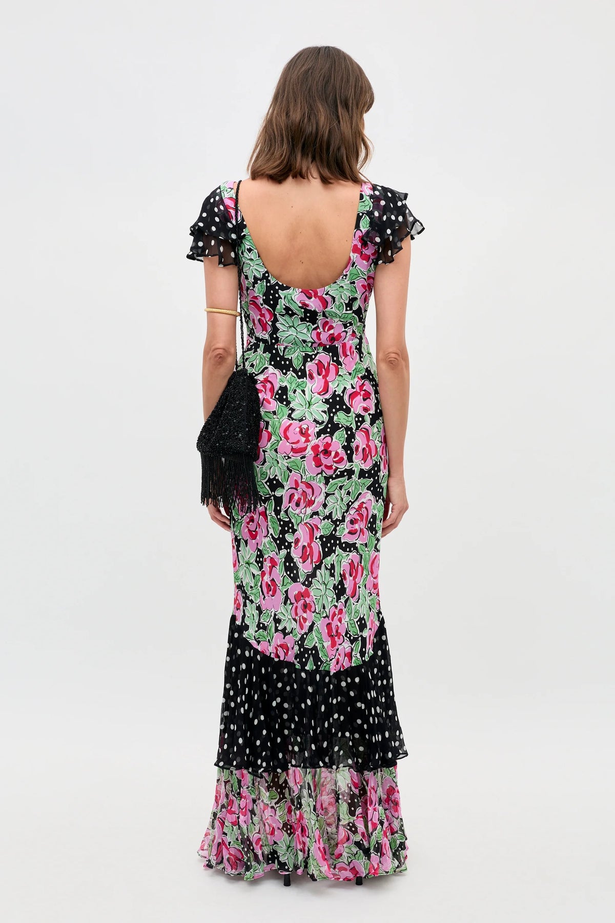 A woman stands facing away, wearing the Ordinia - Rose Dot Mix Black dress—a long style with ruffled sleeves and hem, pink flowers, green leaves, black sections with white polka dots—and carrying a black fringed shoulder bag.