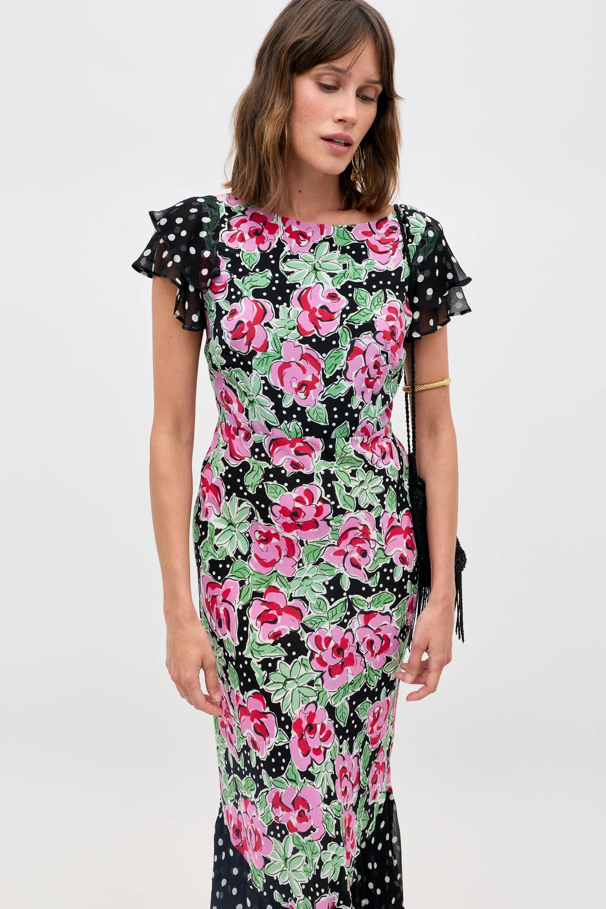 A woman wears the Ordinia - Rose Dot Mix Black dress, featuring a vibrant pink and green floral pattern with white polka dots on black and short ruffled sleeves, standing against a plain light backdrop.