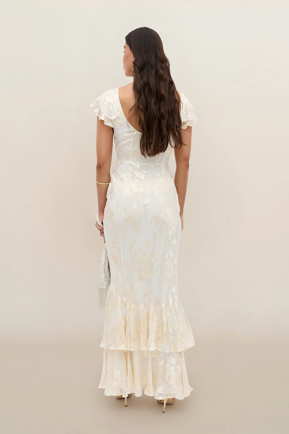 A woman with long wavy hair, seen from behind, wears the Ordinia - Bouquet Devore Cream dress—an elegant off-white floral piece with flutter sleeves and a ruffled hem—holding a small purse against a neutral background.