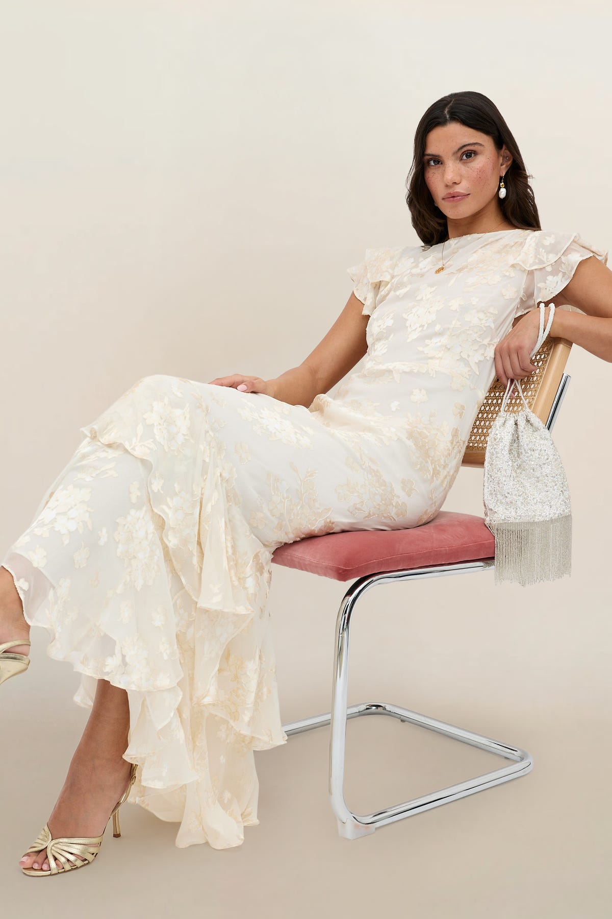 Wearing the Ordinia - Bouquet Devore Cream gown, a woman with shoulder-length dark hair sits on a modern pink-seated chair, holding a beaded purse. She completes her look with gold heels and drop earrings against a neutral background.