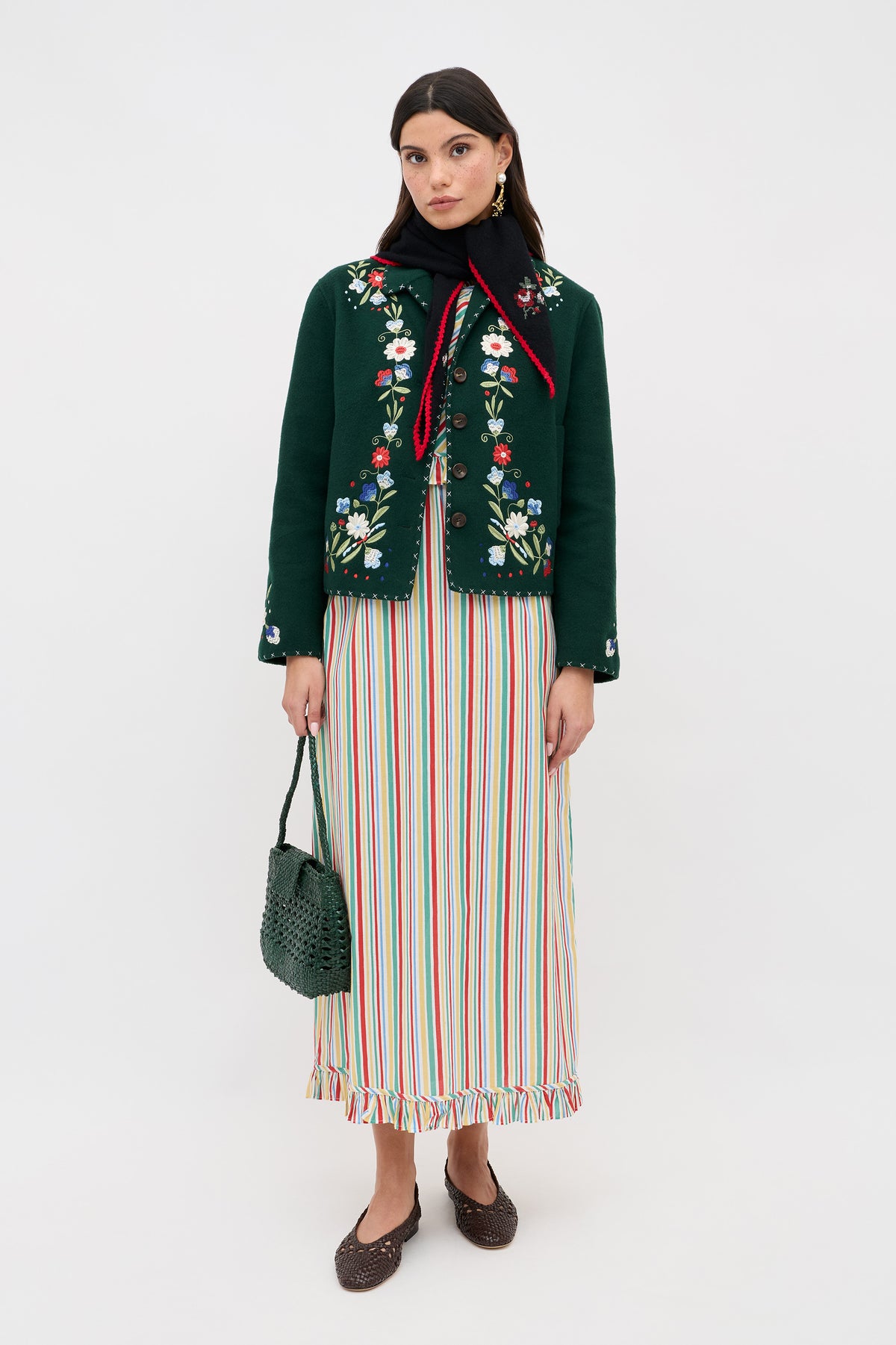 A woman wearing a green embroidered jacket, black scarf with red trim, brown shoes, and holding a green woven handbag stands against a plain white background. She is dressed in the Arabelle - Maxime Stripe Ivory maxi dress.