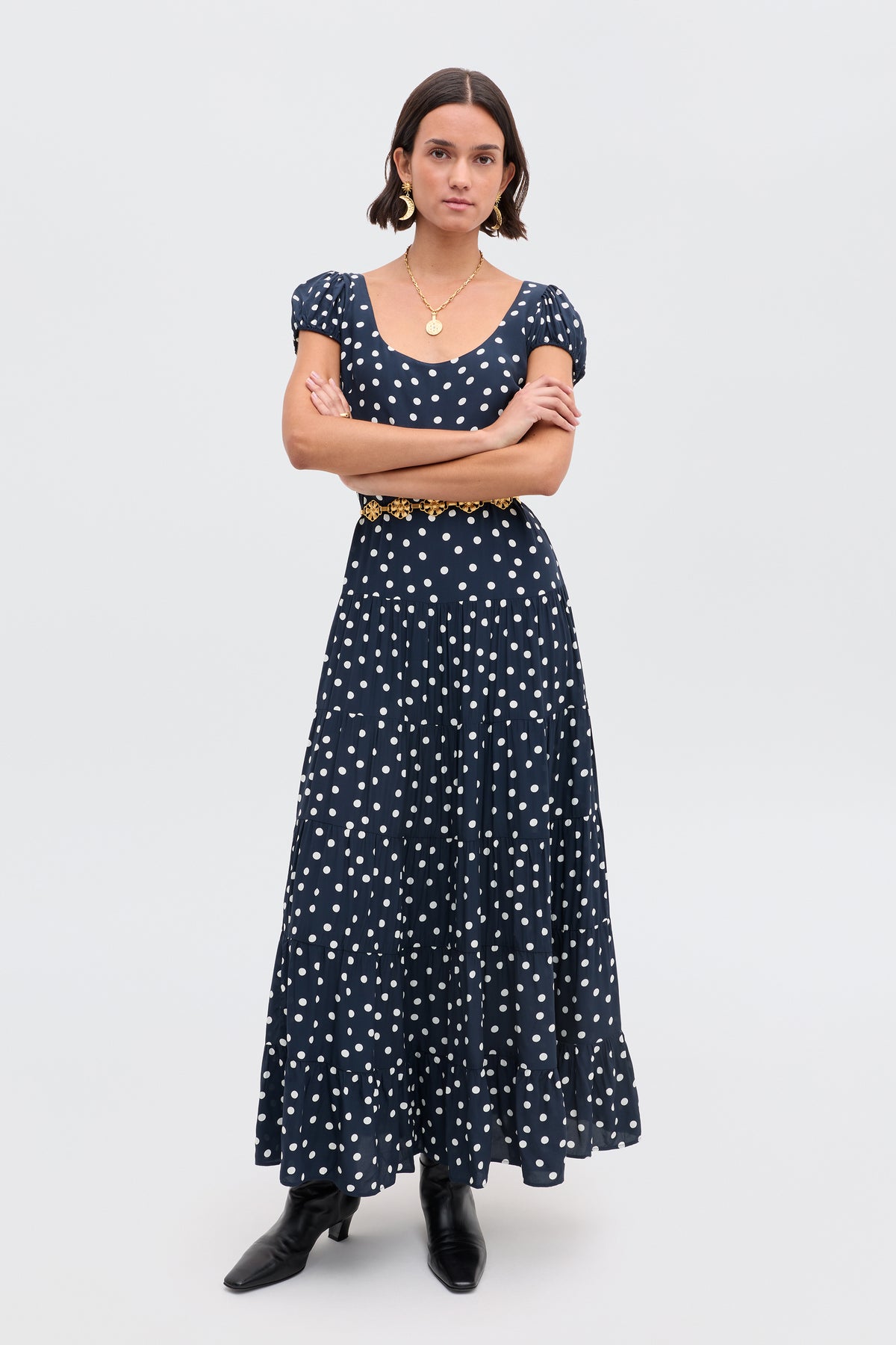 A woman stands with arms crossed, wearing the Remi - Bohemia Spot Deep Navy dress with a black belt and boots, hoop earrings, and a gold necklace against a plain background.