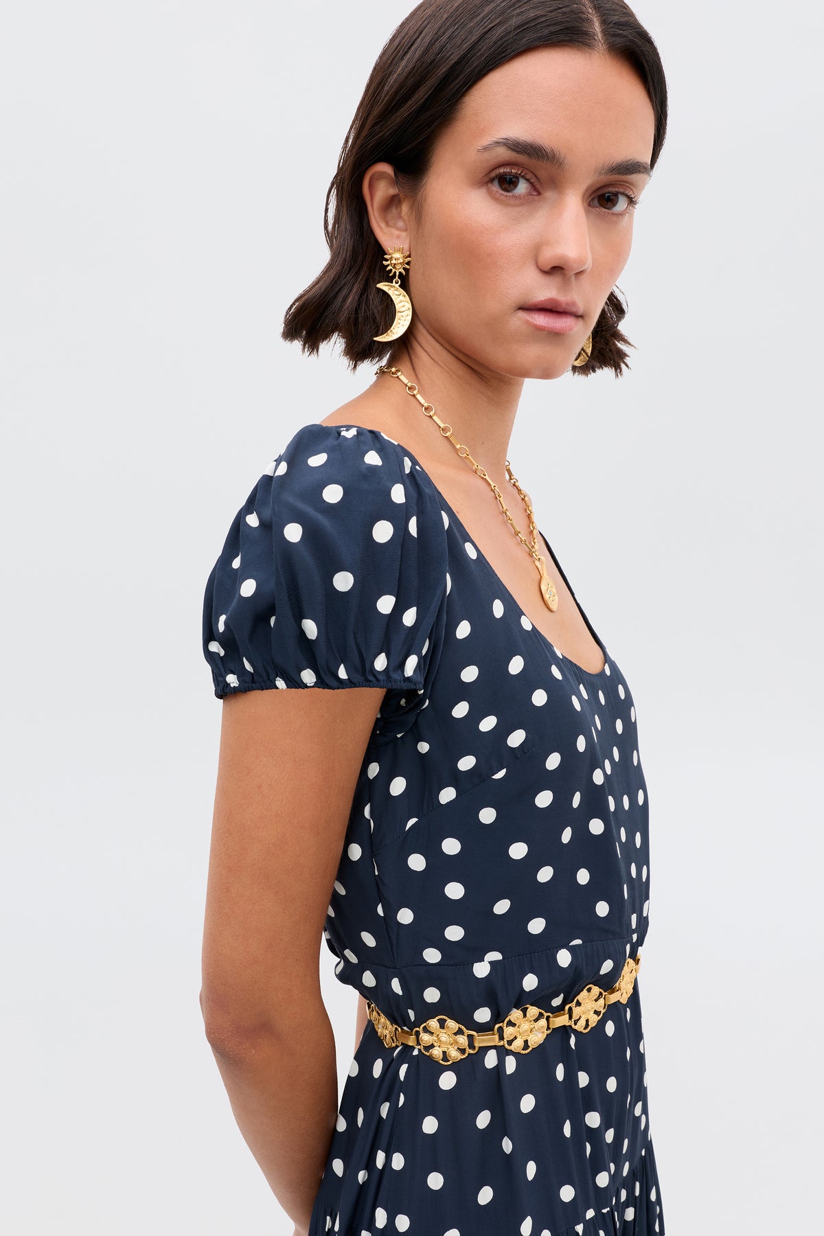 A woman with short dark hair wears the Remi – Bohemia Spot Deep Navy dress, paired with gold earrings, a gold necklace, and a decorative gold belt, standing against a plain light background.