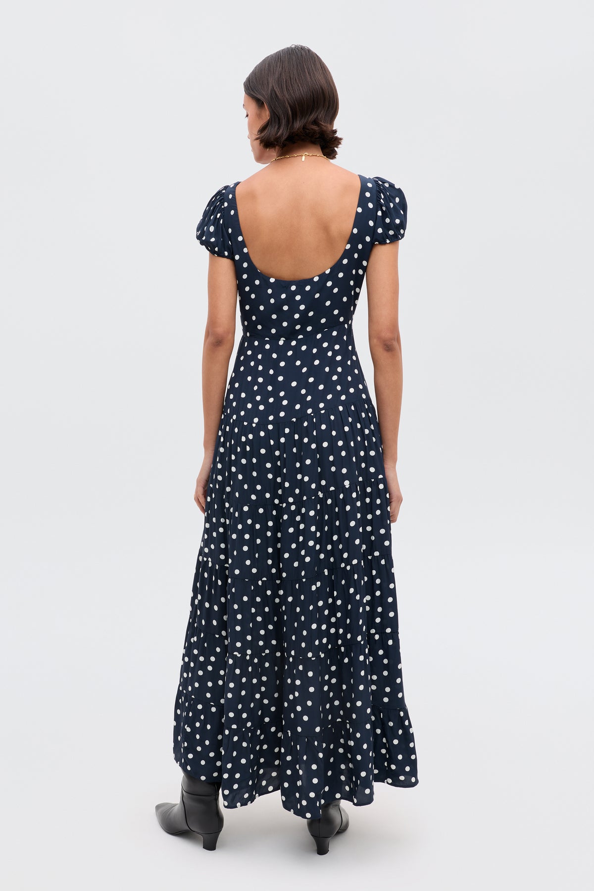 A person with short dark hair, seen from behind, wears the Remi - Bohemia Spot Deep Navy dress and black ankle boots, standing against a plain light background.