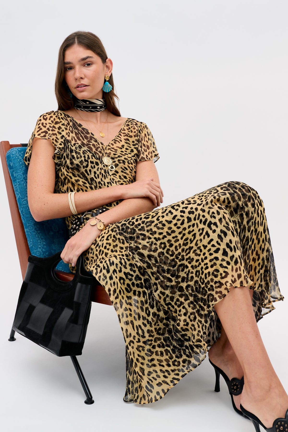 A woman with long brown hair and a calm expression sits on a chair, wearing the Bexley - Nina Leopard Brown dress, black heels, gold jewelry, turquoise earrings, a black scarf around her neck, and holding a black handbag.