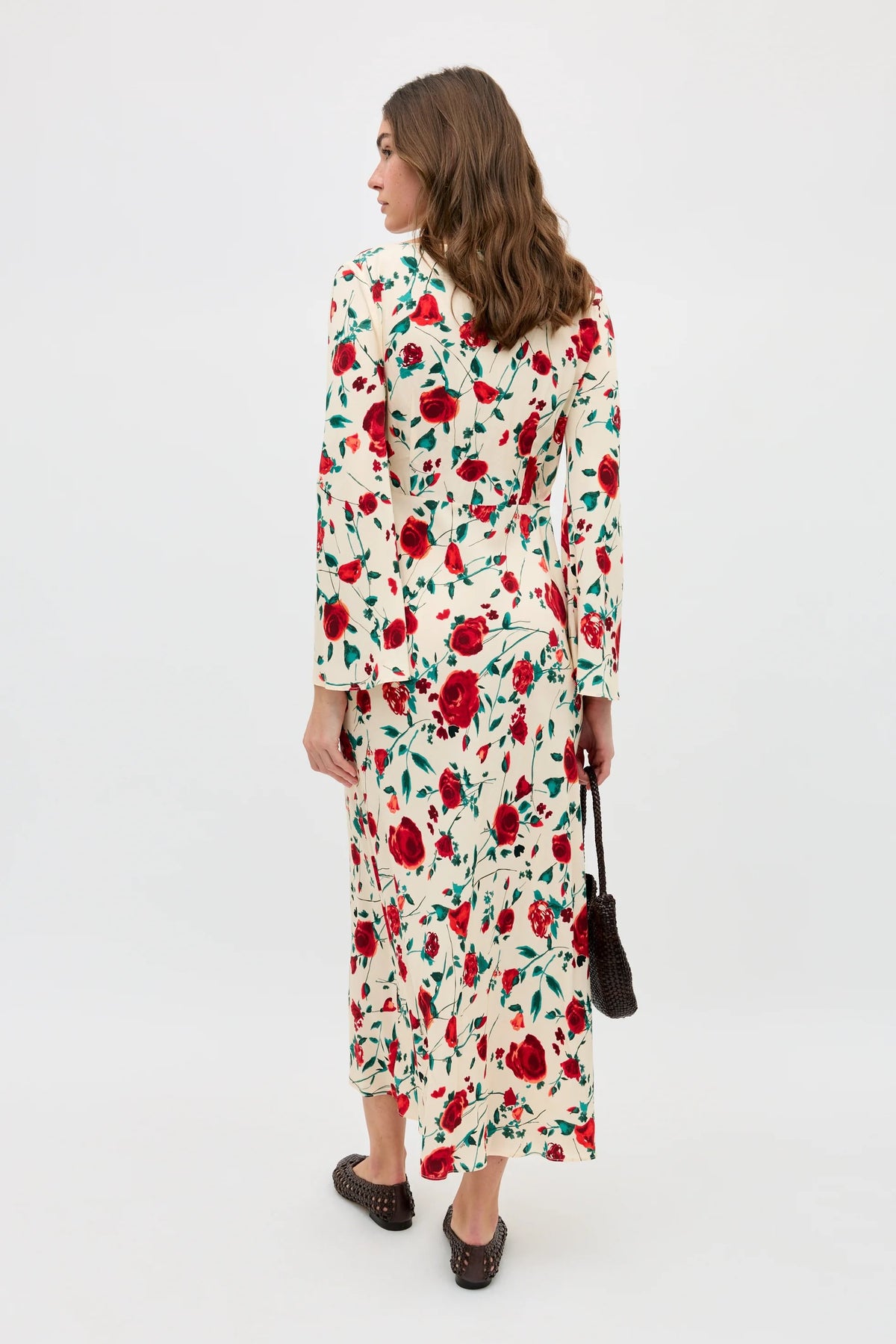 A woman with wavy brown hair stands facing away, wearing the Gwenyth - Claudia Rose Cream dress—an ankle-length, long-sleeved cream dress with a red and green floral pattern—paired with black shoes and holding a small black handbag.