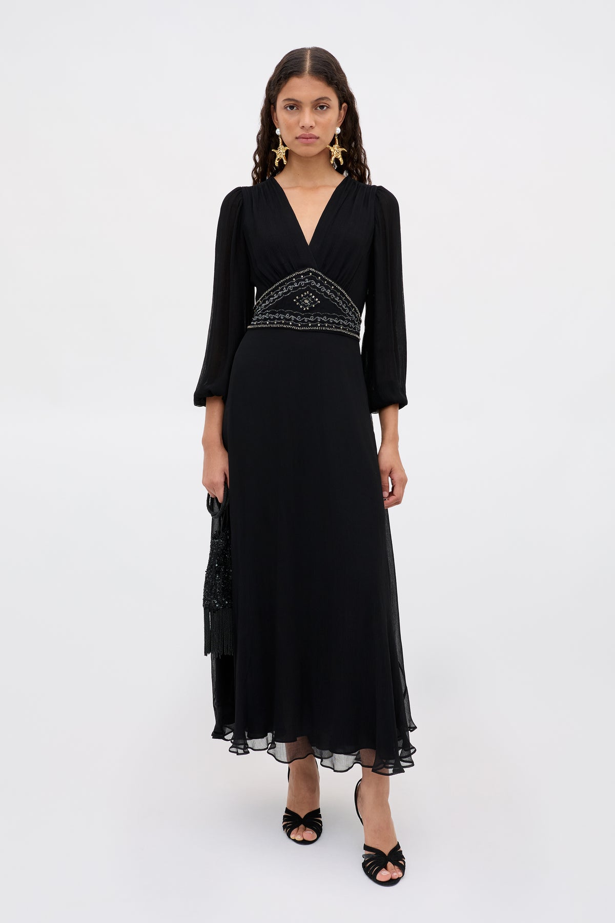 A woman stands against a white background, wearing the Kaesha - Diana Embellishment Black long v-neck dress with three-quarter sleeves and a beaded waistband, paired with black open-toe heels, and holding a black bag in her left hand.
