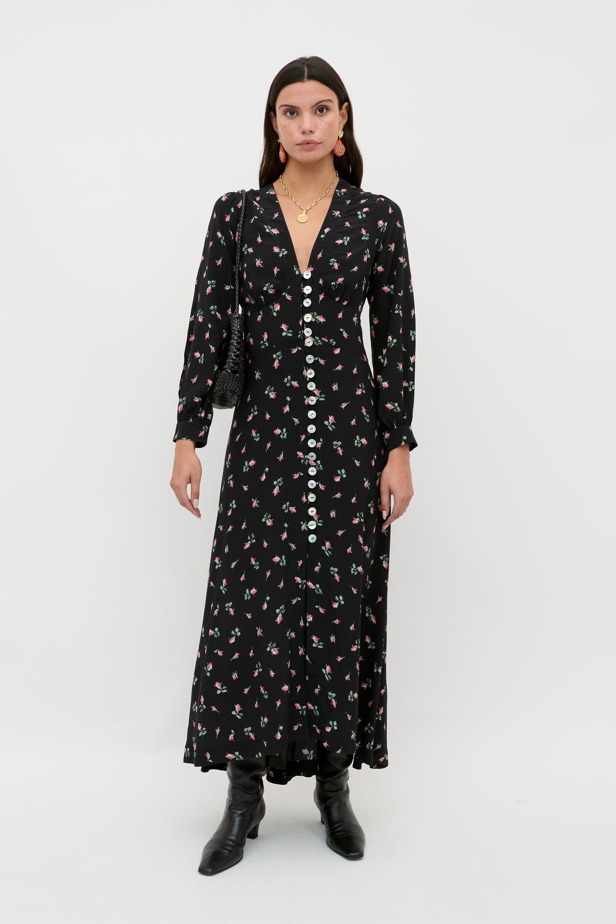 A woman wears the Avalyn - Sienna Bud Black long sleeve midi dress with vintage-inspired cuffs, paired with black boots, a small black purse, and gold jewelry, standing against a plain white background.