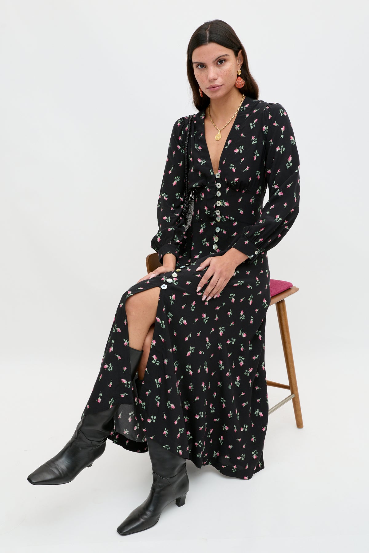 A woman wearing the Avalyn - Sienna Bud Black long sleeve midi dress with vintage cuffs, button-down front, and a thigh-high slit sits on a wooden chair in black boots and gold jewelry, posing against a plain white background.
