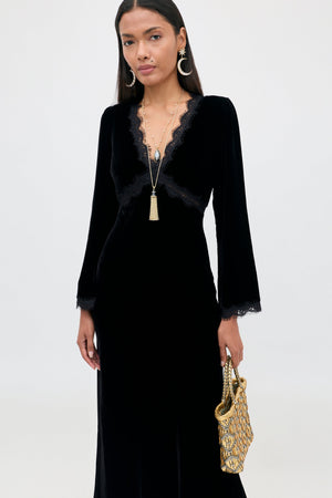 A model wears the Alessi - Black long-sleeved velvet dress with a V-neckline, lace detailing, and a gold-toned metallic bag.