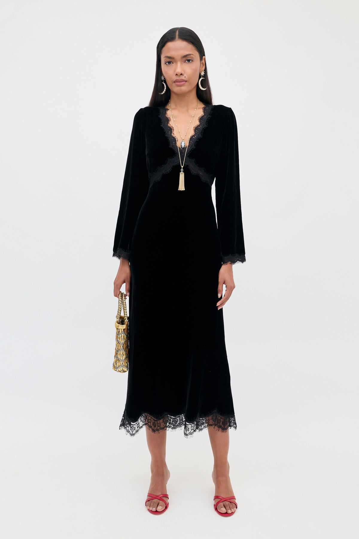 A person wearing the Alessi - Black midi-length velvet dress with a V-neckline, long sleeves, and boho-glamour lace trim at the collar, cuffs, and hem, holding a gold handbag.