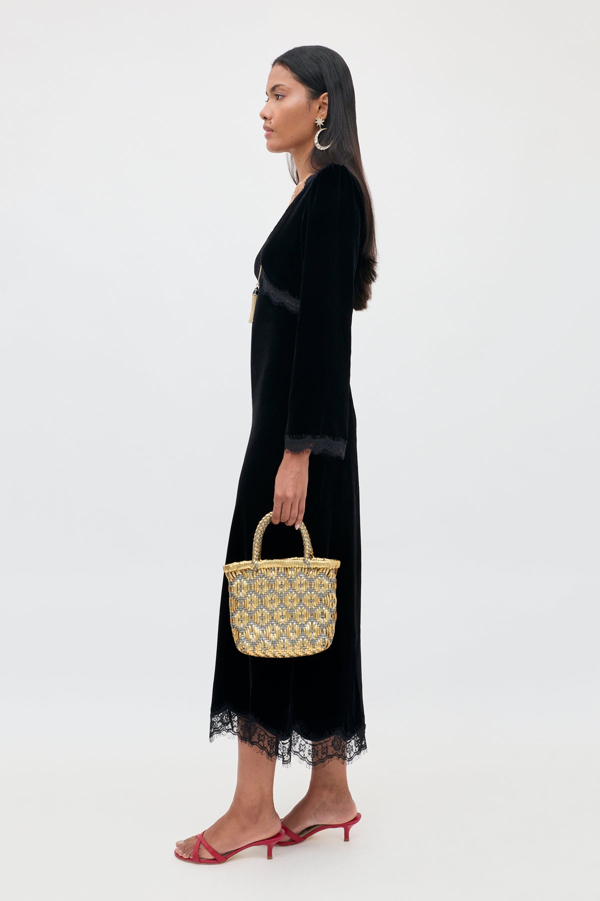 A woman in profile wears an Alessi - Black long-sleeved velvet midi dress with lace detailing and a boho-glamour aesthetic, paired with a small metallic gold woven basket bag and red heeled sandals.