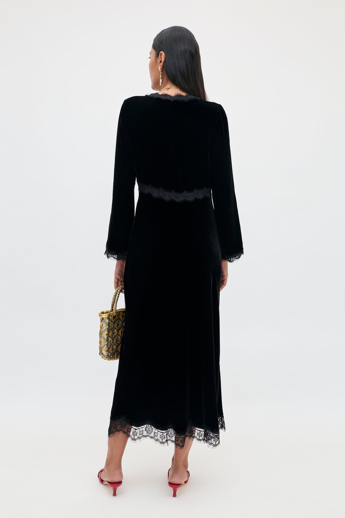 Seen from behind, a person wears the Alessi - Black long-sleeved midi dress with boho-glamour lace detailing at the waist, cuffs, and hem, paired with red slide heels and holding a small woven handbag.