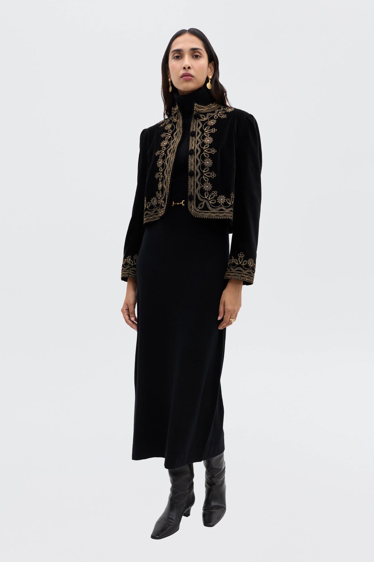 A woman with long dark hair poses against a plain white background, wearing the Elliette - Black minimalist long black knitted dress, paired with black boots and a black cropped jacket featuring gold embroidery.