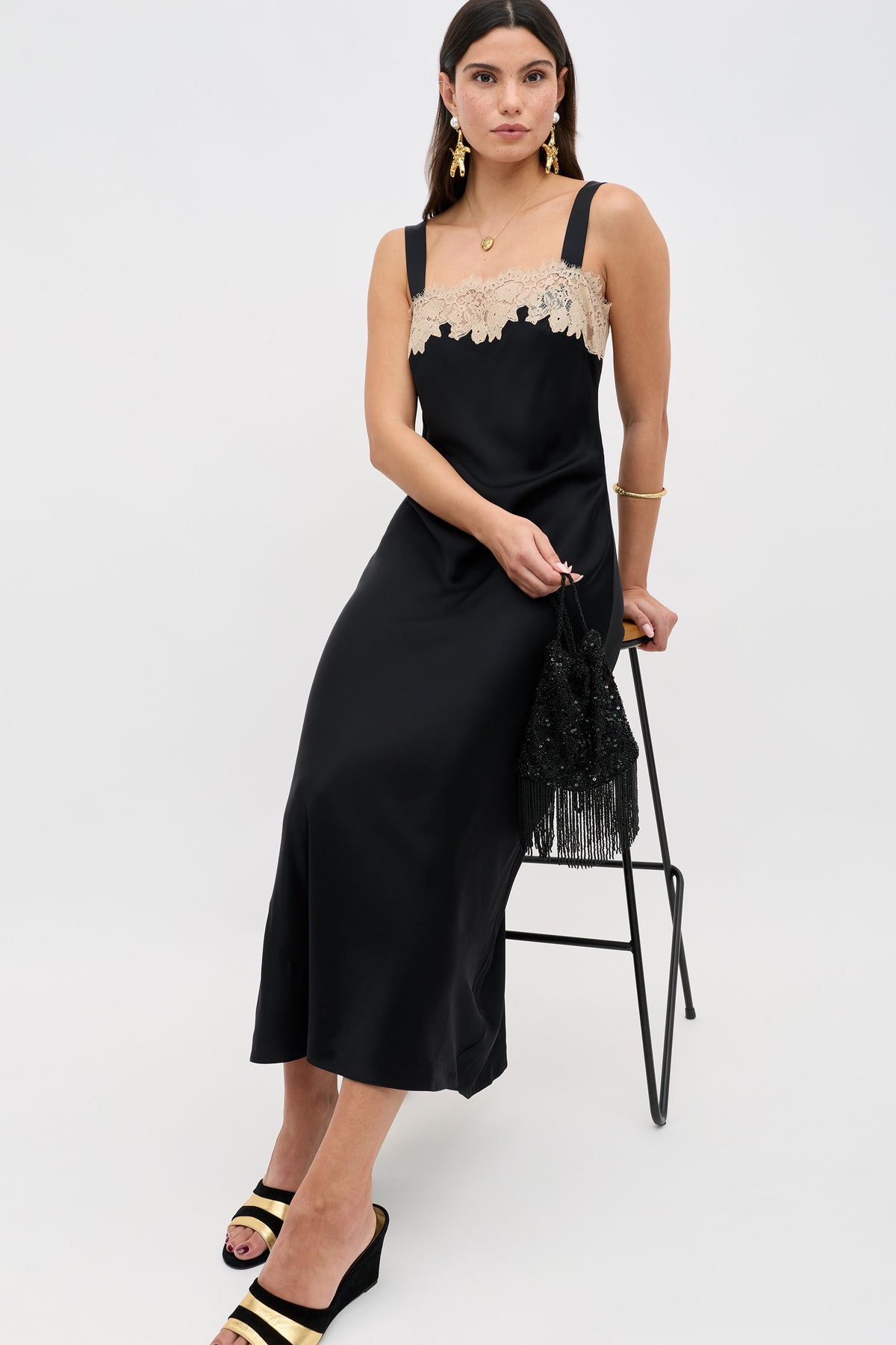 A woman in the Rosamarie - Black sleeveless midi dress with beige lace at the neckline sits on a stool, styled with gold jewelry, black and gold platform sandals, and a black fringed beaded bag.