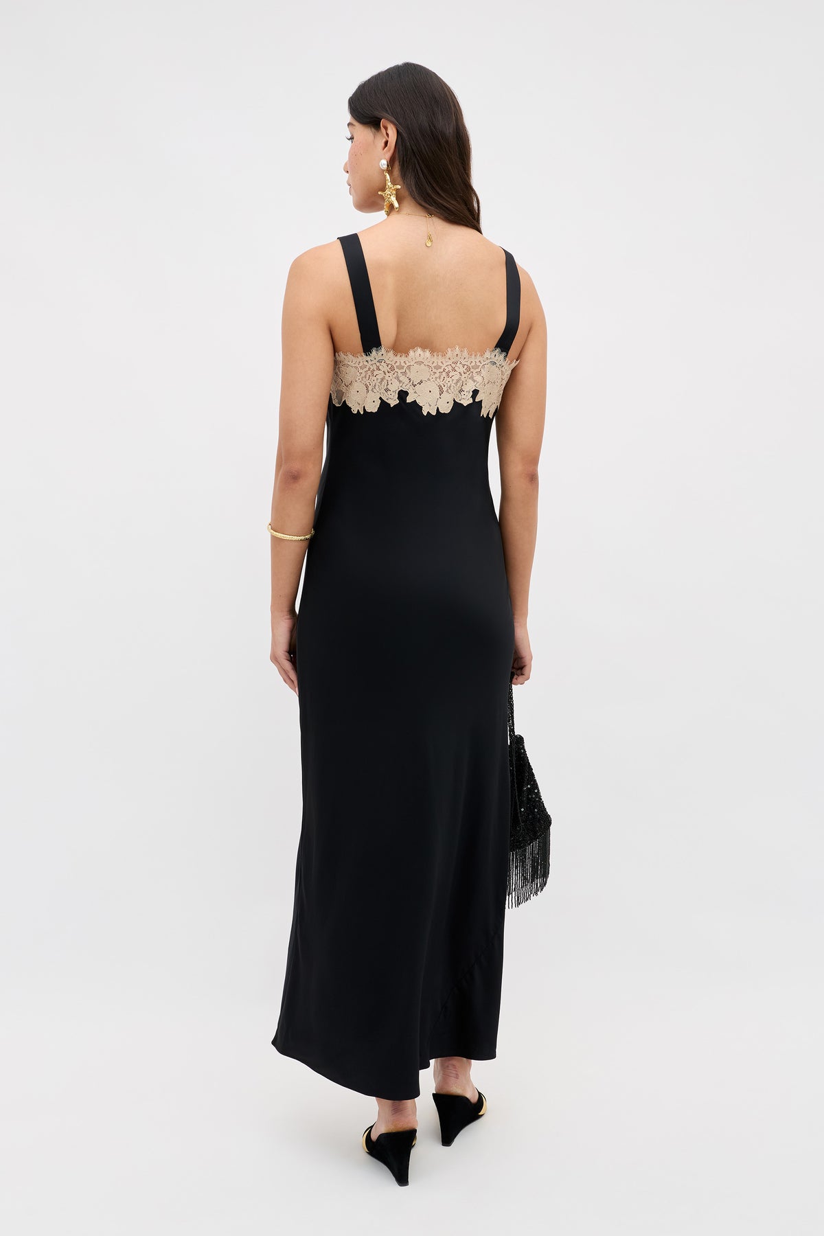 Wearing the Rosamarie - Black sleeveless gown with beige lace back, a woman faces away holding a black fringed bag. She has long dark hair down and accessorizes with gold earrings, a bracelet, and black pointed shoes.