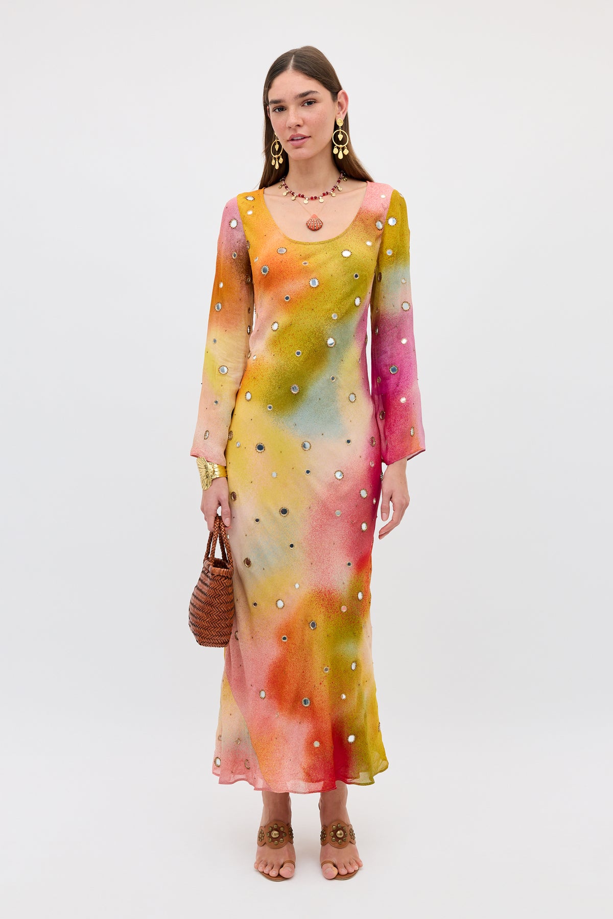 A woman wearing the Noraya - Jessica Alexandra Multi dress, a long, multicolored Liberty fabric piece with long sleeves and delicate embellishments, holds a brown woven bag, statement earrings, and sandals as she stands against a plain white background.