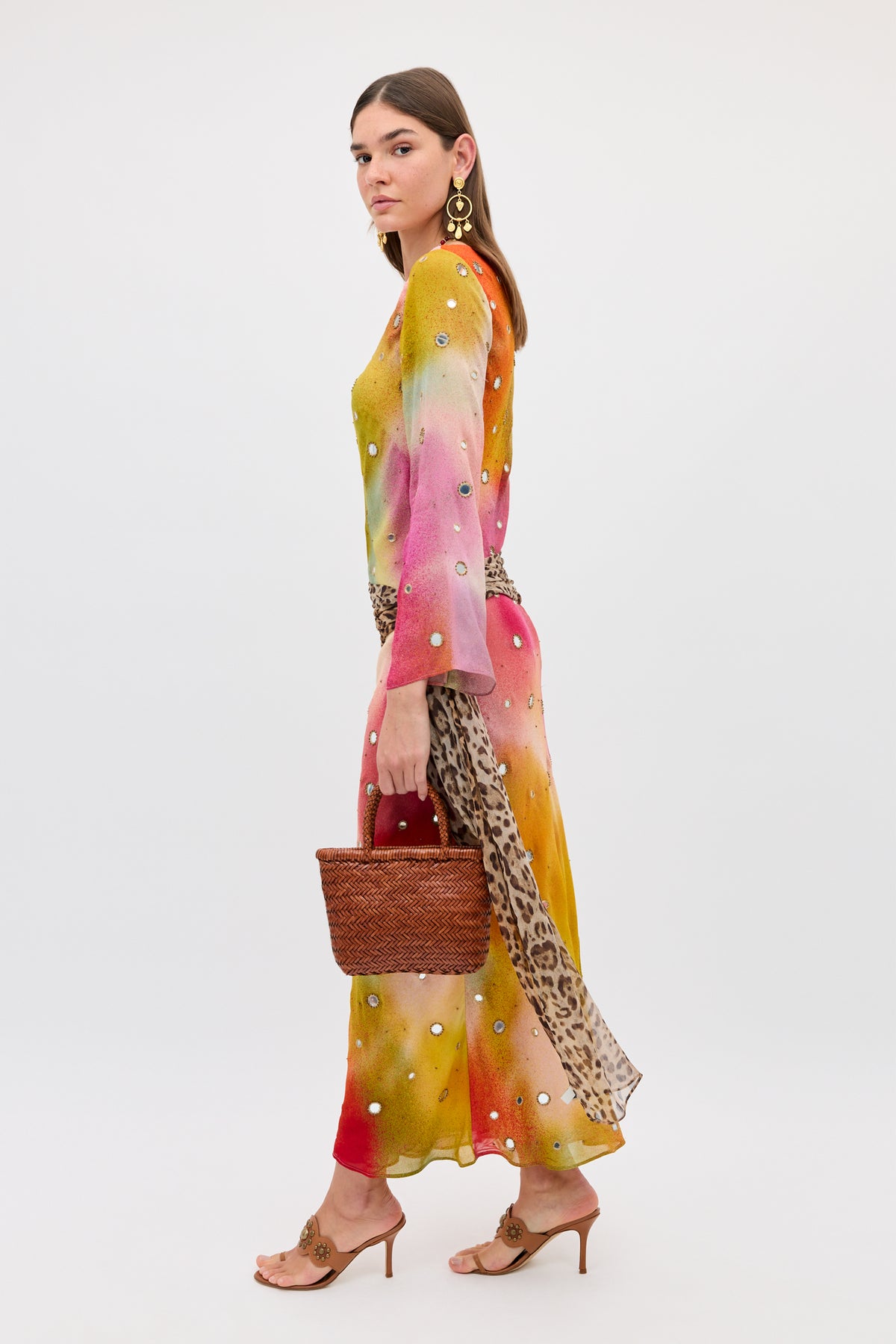 A woman stands against a plain background in the Noraya - Jessica Alexandra Multi, a colorful, long-sleeved silk dress with pearl-like accents. She holds a woven brown bag and accessorizes with heeled sandals, gold earrings, and a leopard print belt. Her straight hair completes the look.