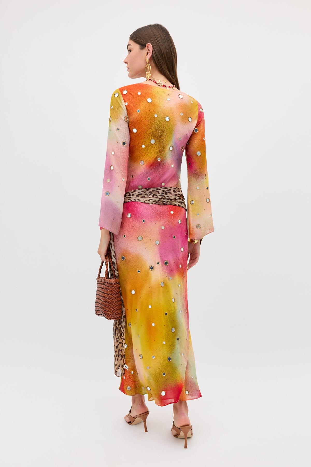 A woman wearing the Noraya - Jessica Alexandra Multi, a long, brightly colored silk dress with polka dots, stands with her back to the camera. She carries a woven bag, wears heeled sandals and large earrings, and has straight brown hair.