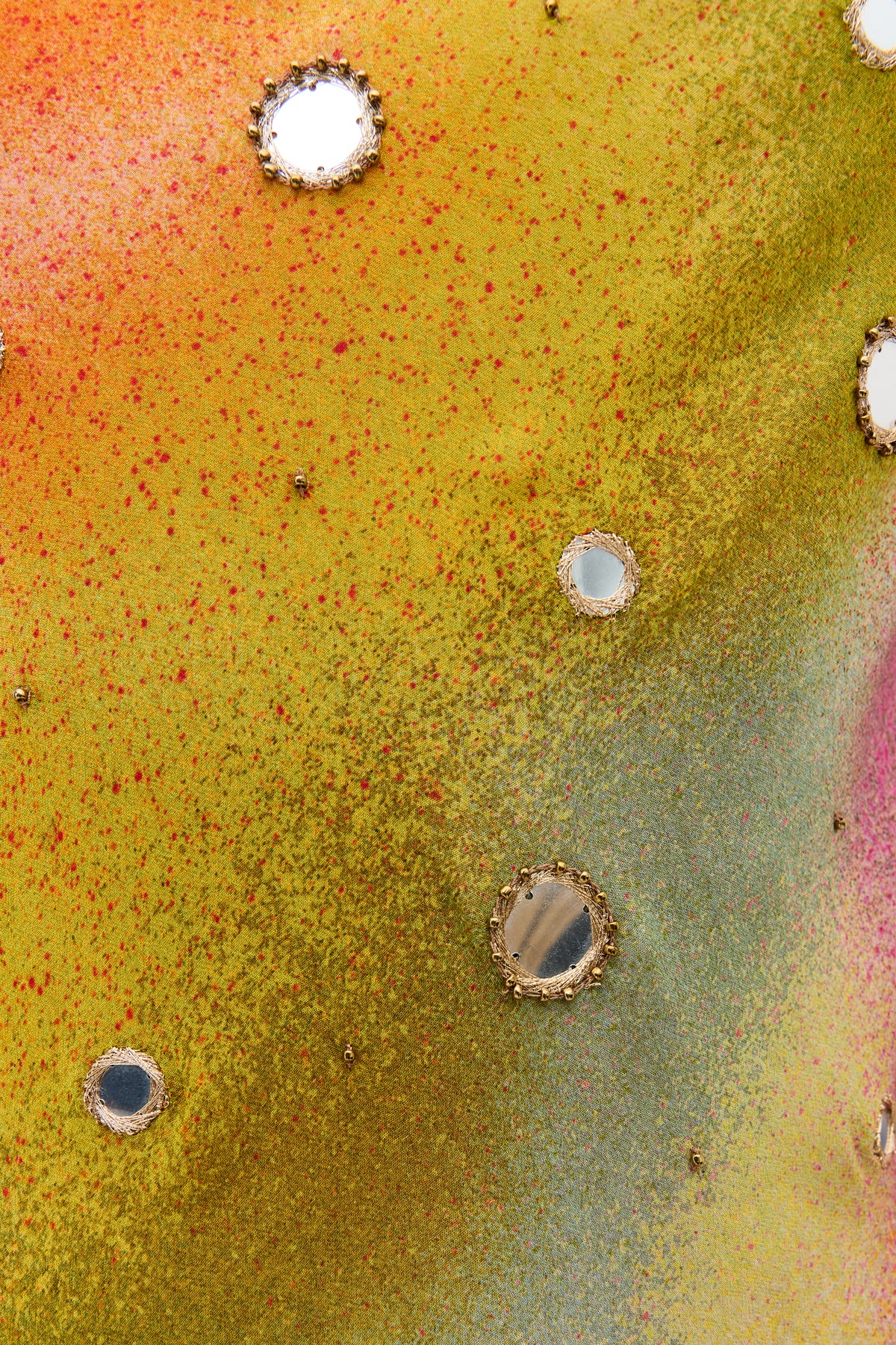 A colorful textured surface with shades of yellow, green, and red, reminiscent of a vibrant Liberty fabric, featuring small round mirrors bordered by metallic edges scattered across the image—introducing the Noraya - Jessica Alexandra Multi.