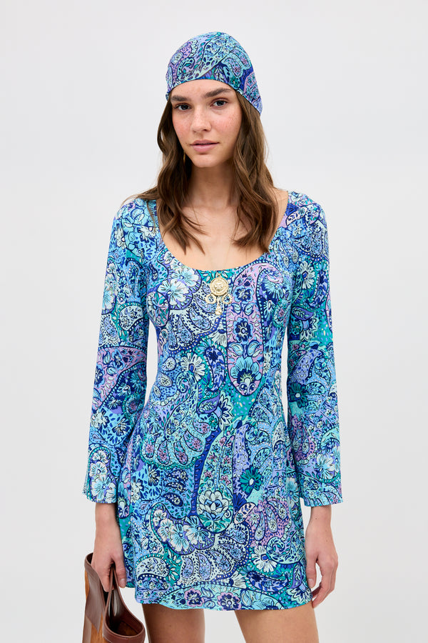 A woman wears the Dulce - Paisley Floral Blue dress with a matching headscarf, standing against a plain background and holding a brown handbag by her side.
