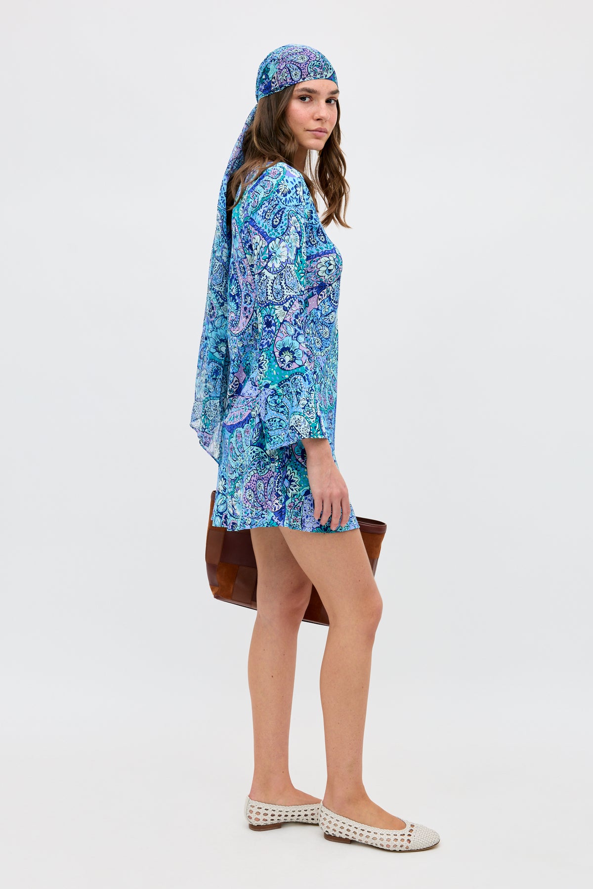 A woman stands sideways against a plain white background, wearing the "Dulce - Paisley Floral Blue" dress with a matching headscarf. She carries a brown bag, wears white woven flats, and looks toward the camera.