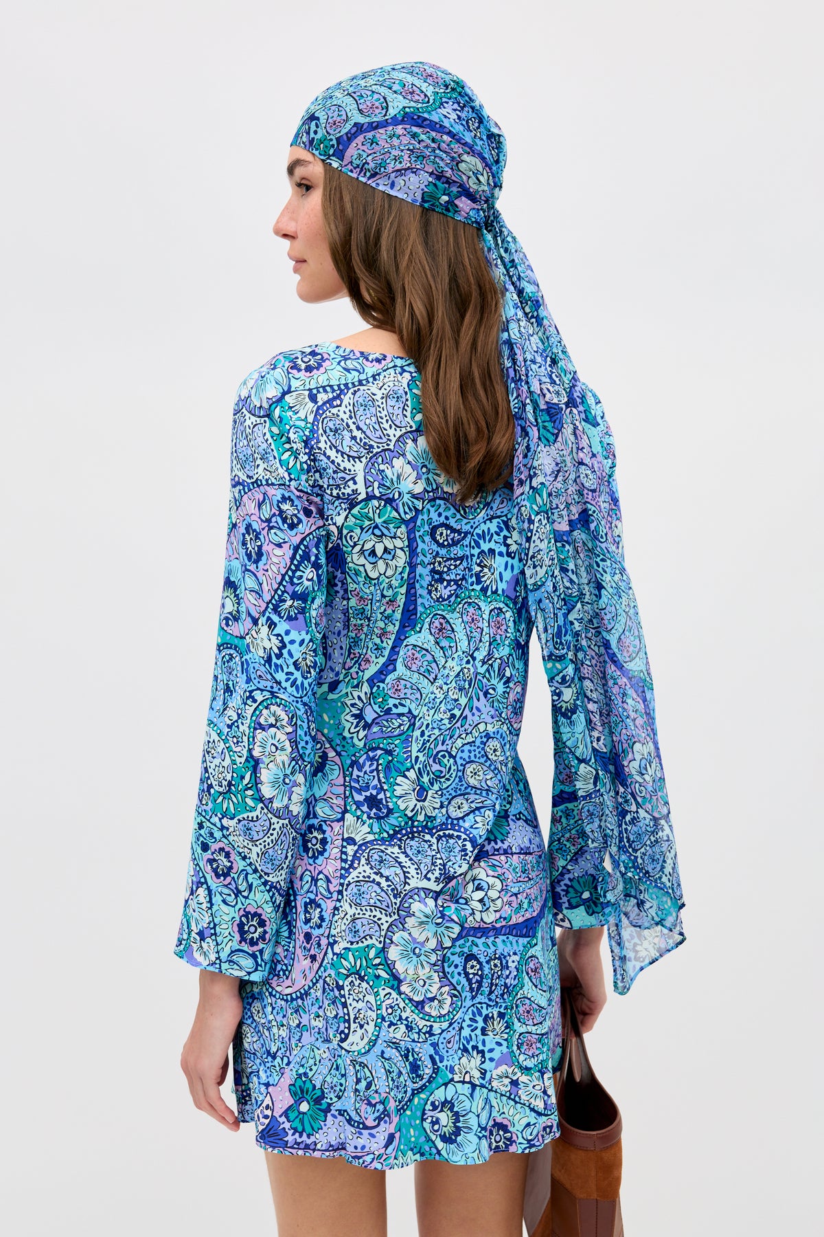 A woman wearing the "Dulce - Paisley Floral Blue" dress with a matching headscarf stands facing away, holding a brown bag, against a plain light background.