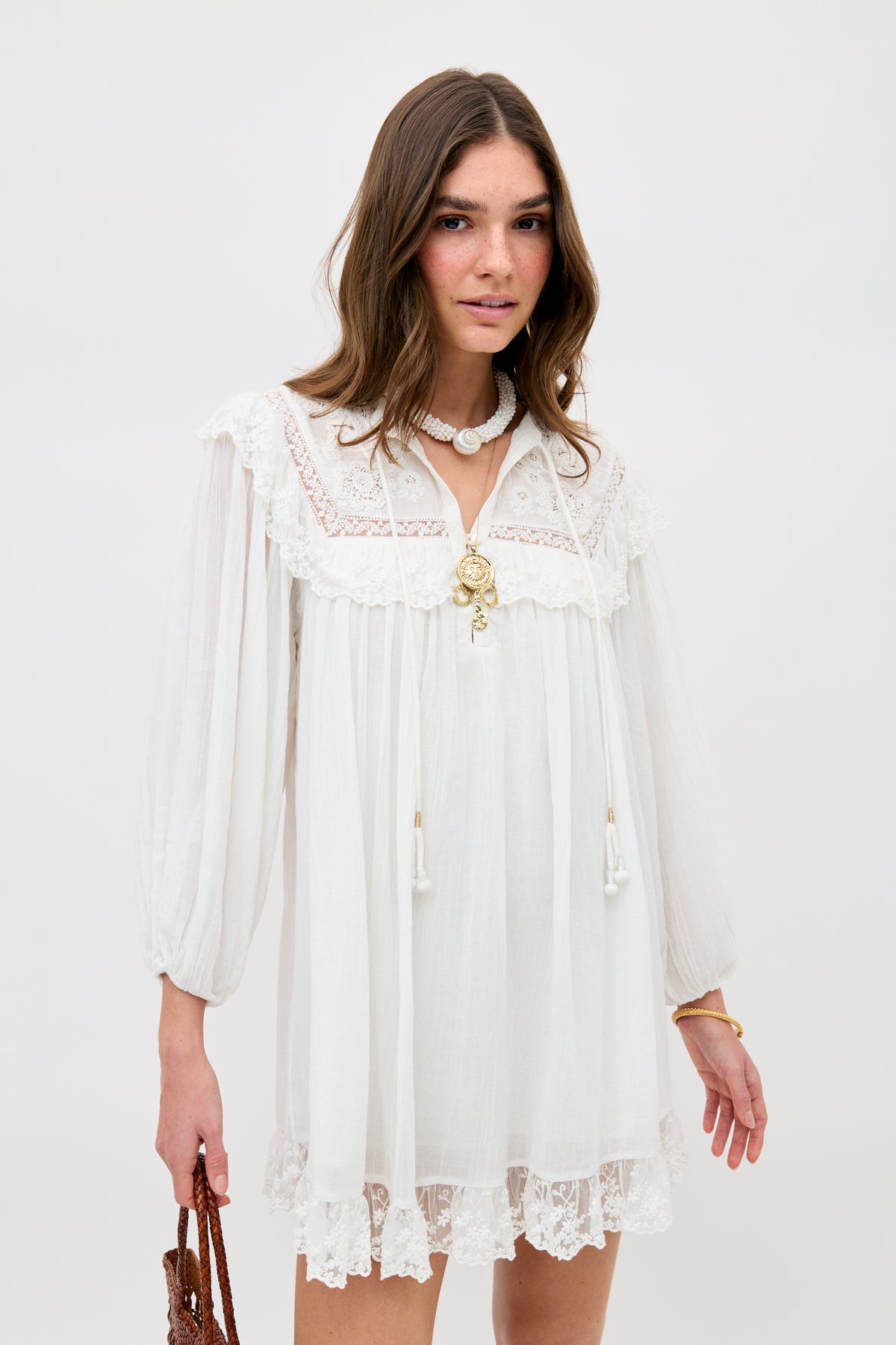 A woman in the Inez - Lilly Embroidery Ivory dress, featuring loose long sleeves and lace trim, accessorized with layered necklaces and a brown woven bag, stands against a plain background.