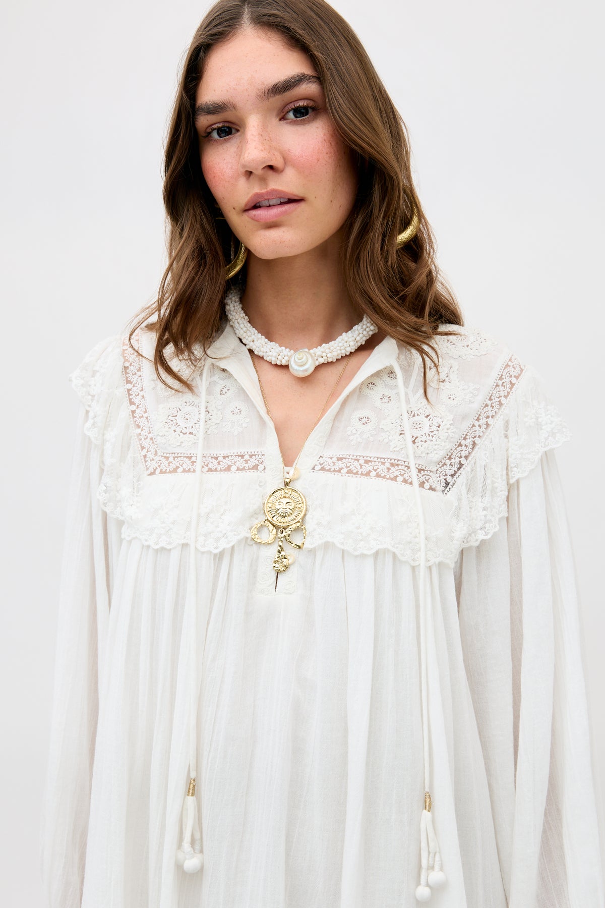 A woman with long brown hair wears the Inez - Lilly Embroidery Ivory dress, featuring lace details and tassels, paired with hoop earrings and layered gold and pearl necklaces, standing against a light background.
