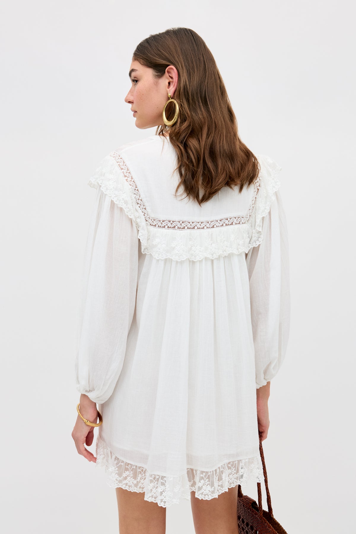 A woman with wavy brown hair wears the Inez - Lilly Embroidery Ivory long-sleeved dress, accessorized with large gold hoop earrings, a gold bracelet, and a brown woven bag. She stands facing away against a plain white background.