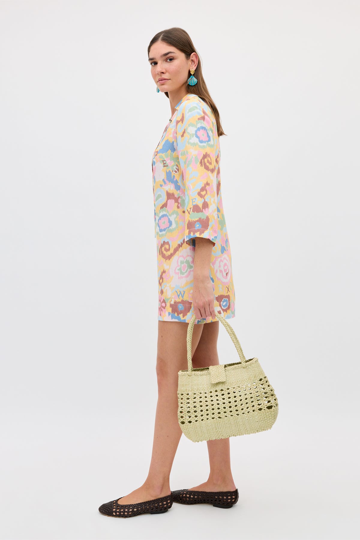A woman with long brown hair stands against a plain white background, wearing a pastel floral dress and black flats. She holds the Dragon Diffusion Niamh - Butter vintage woven bag and accessorizes with turquoise earrings.