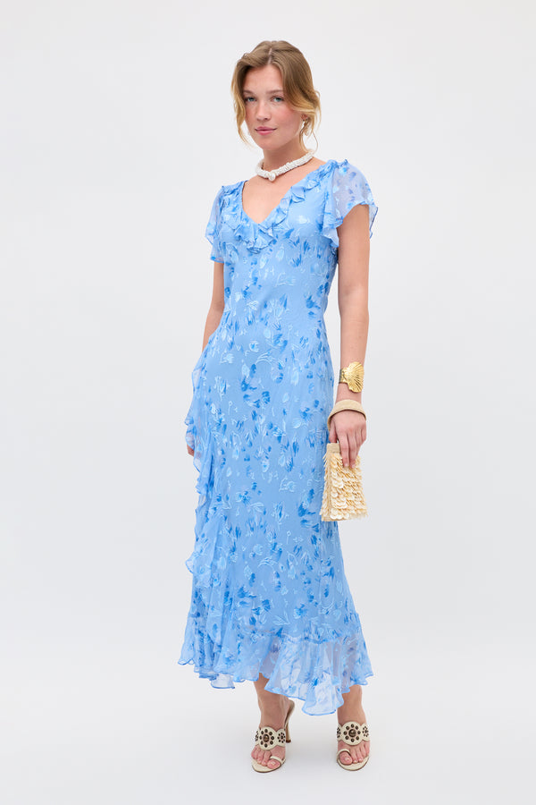 A woman models the Meadow - Ava Devore Blue dress, a light blue, floral, ruffled midi dress with short sleeves. She accessorizes with a gold textured clutch, white sandals, a shell necklace, and a gold cuff bracelet against a plain white background.