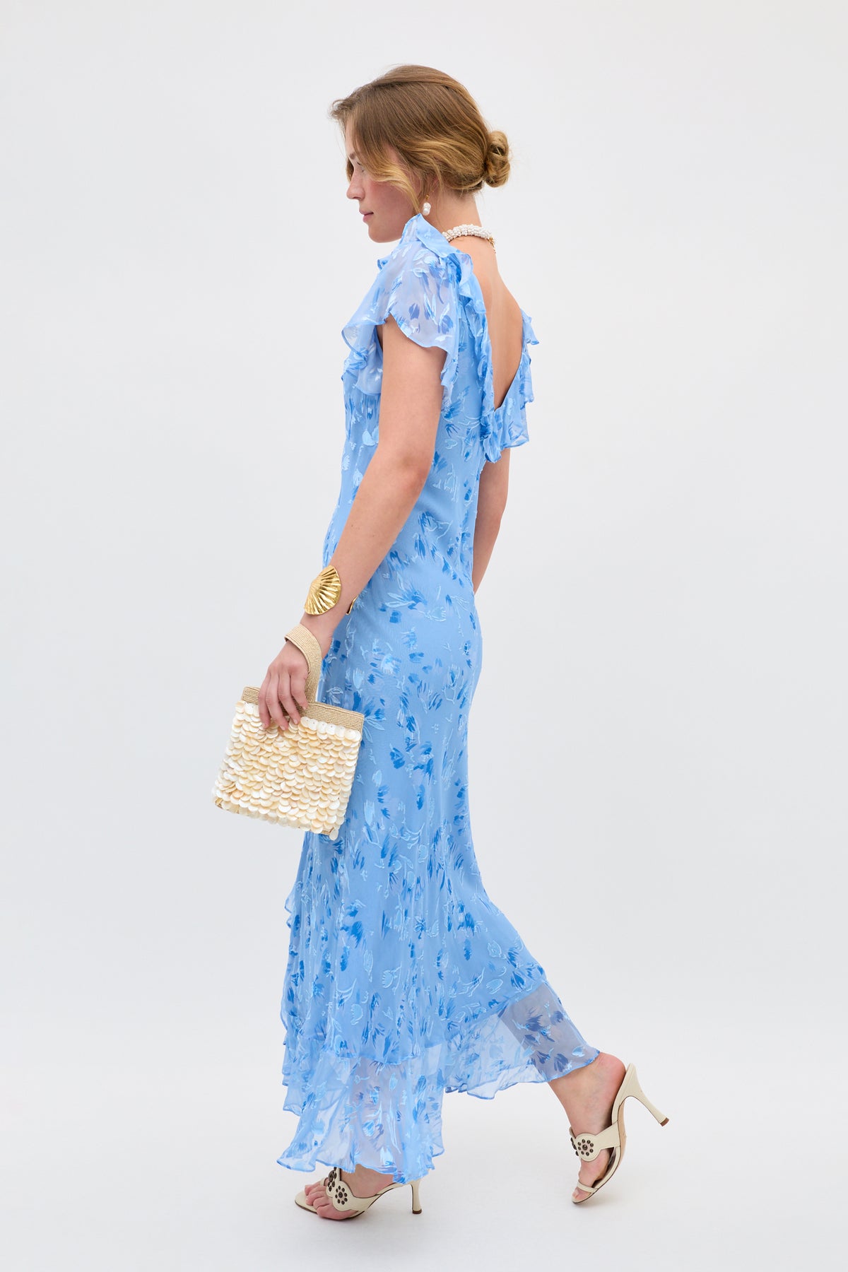 A woman in the Meadow - Ava Devore Blue dress, featuring light blue florals and ruffle sleeves, stands against a plain light background, holding a woven handbag and accessorized with white heels and gold jewelry, facing to the side.
