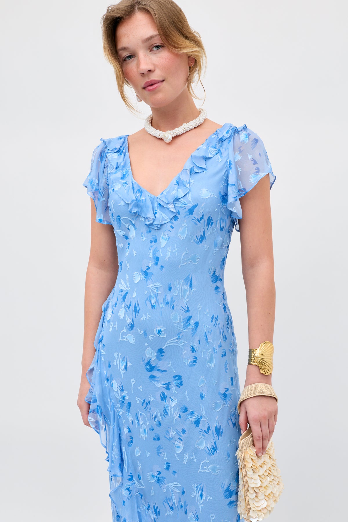 Wearing the Meadow - Ava Devore Blue dress, a woman stands against a plain background, styled with a white shell necklace, gold bracelet, and a cream-colored textured clutch.