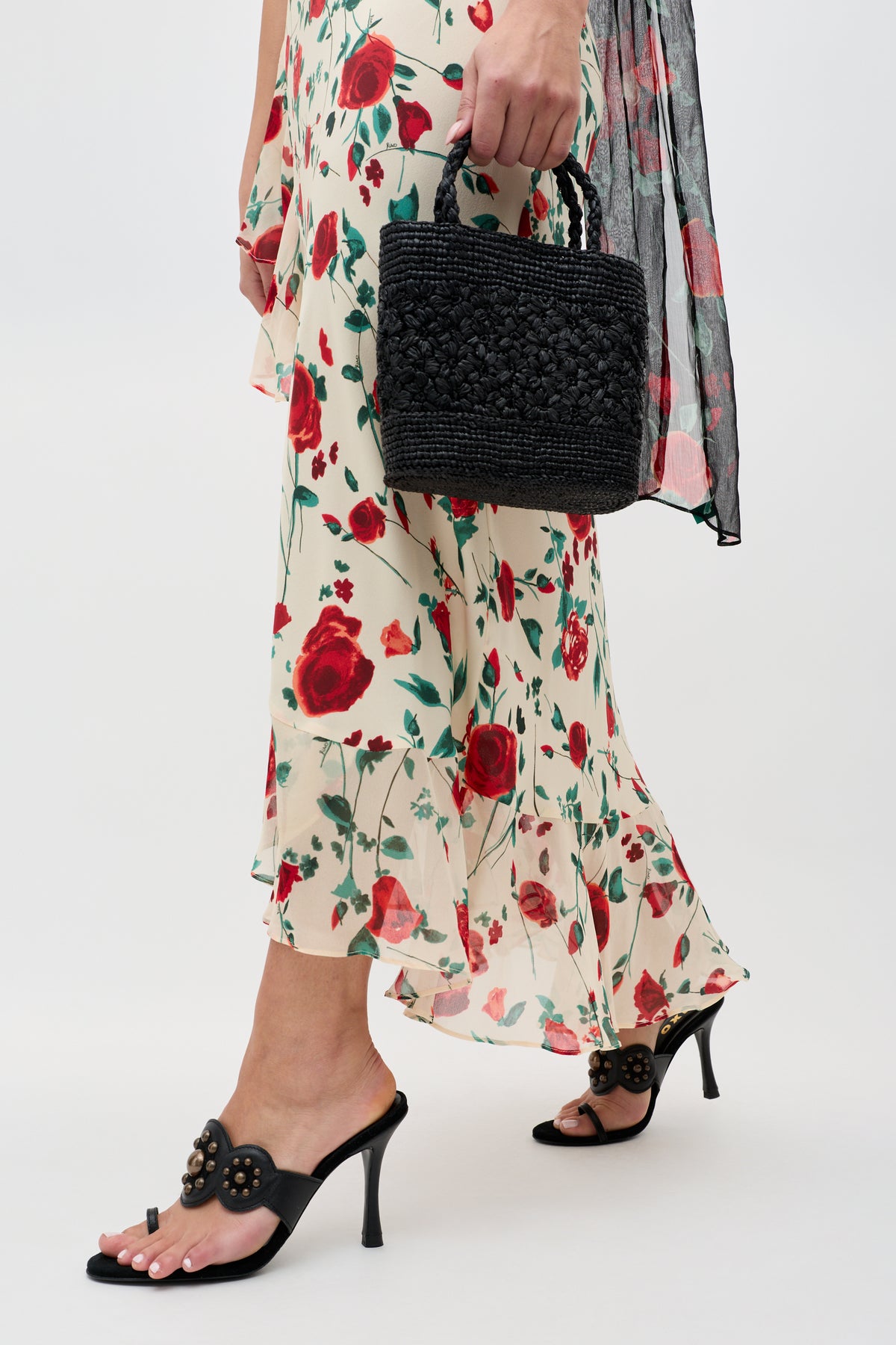 A person wearing the Meadow - Claudia Rose Cream dress with red rose prints and ruffle hem, black high heel sandals, and holding a small black woven handbag; only the lower body and hand are visible.