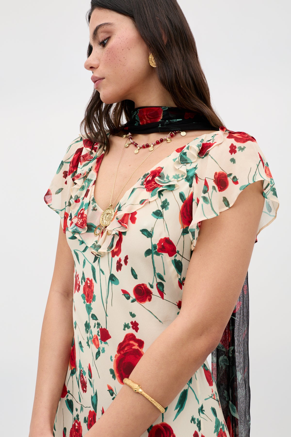 A woman wearing the Meadow - Claudia Rose Cream dress with red floral patterns, gold jewelry, and a sheer black scarf stands with arms crossed and looks downward against a plain light background.