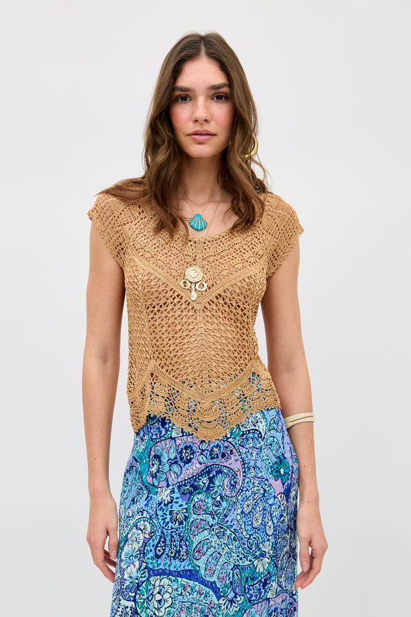 A woman with wavy brown hair wears the Kimbra - Gold necklace layered with other pieces, paired with a tan crochet top and a blue patterned skirt. She stands against a plain light background, looking at the camera with a neutral expression.