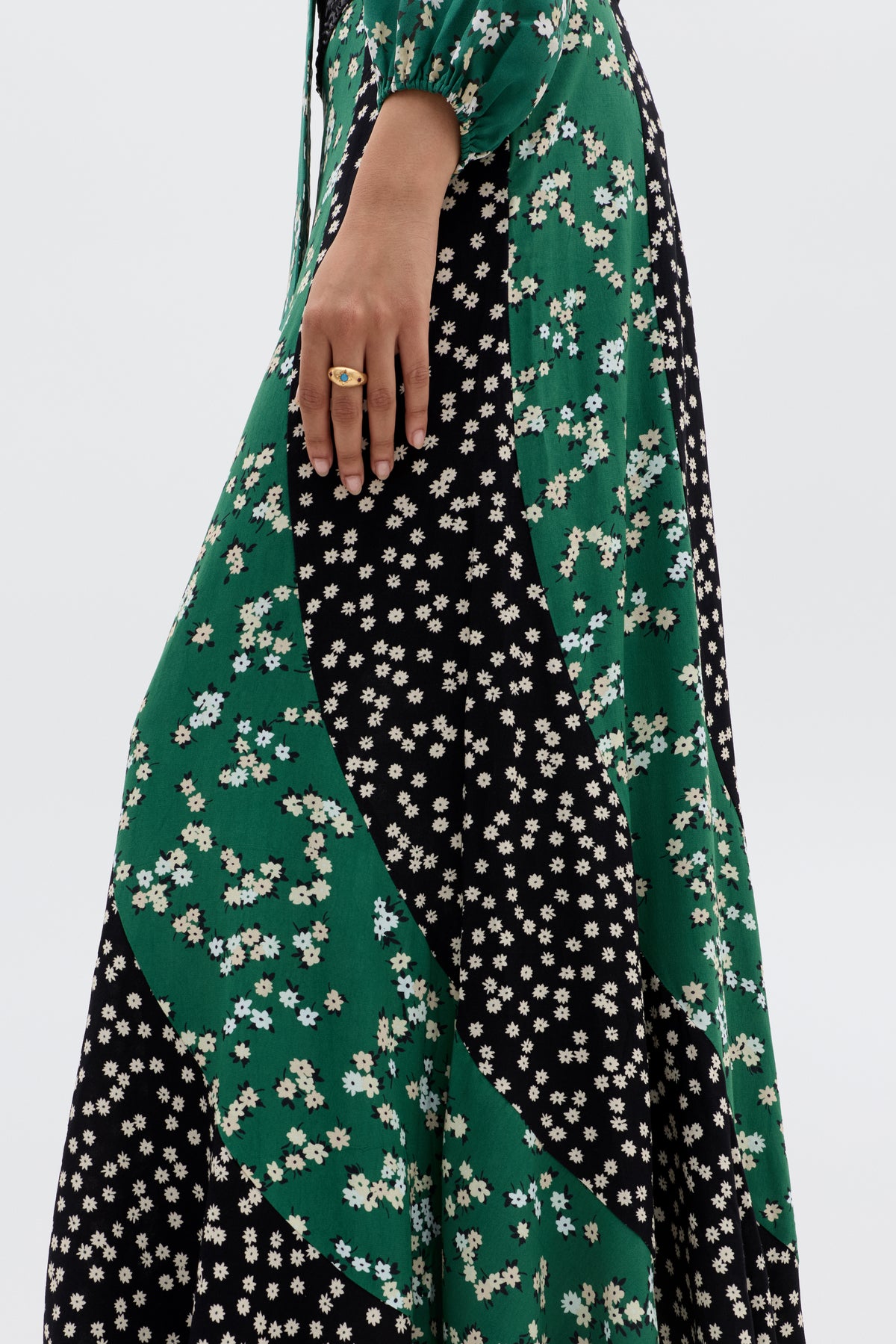 A person models transitional dressing in the Gracella - City Floral Mix Olive, a long dress with green and black panels and winter floral patterns. They wear a gold ring, with their hand resting by their side.