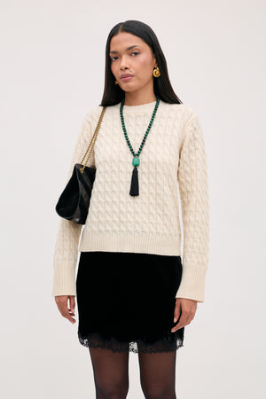 A woman wears a cream cable-knit sweater, a Kornelia - Black velvet mini skirt with a lace hem, and a black shoulder bag with a gold chain strap.