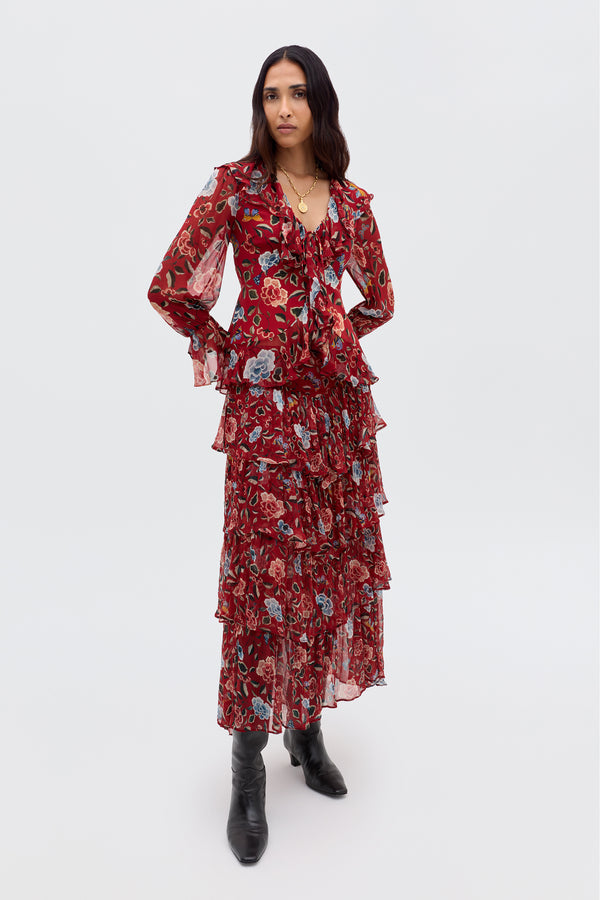 A woman with long dark hair wears the Taryn - Butterfly Floral Burgundy dress, featuring ruffles, long sleeves, and a prairie skirt. She pairs it with black boots and a gold necklace, standing against a plain background.